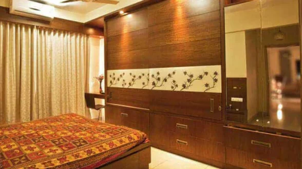 A bedroom with a brown wardrobe, a dressing table with a mirror, and flowery wall patterns.